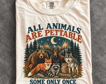 May include: An ivory t-shirt featuring a graphic of a bear, moose, fox, raccoon, owl, and wolf in a forest under a crescent moon. The text reads ALL ANIMALS ARE PETTABLE SOME ONLY ONCE. The shirt has a Comfort Colors brand label at the neck.