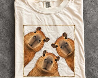 May include: An ivory-coloured t-shirt featuring a square graphic print of three brown capybaras looking directly forward. The shirt has a crew neckline and a Comfort Colors brand label printed on the inside of the collar area.