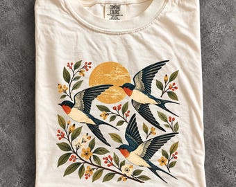 May include: An ivory t-shirt featuring a graphic print of three swallows in flight, surrounded by green leaves, small yellow flowers, and red berries, with a large textured yellow sun positioned behind the birds in the centre.
