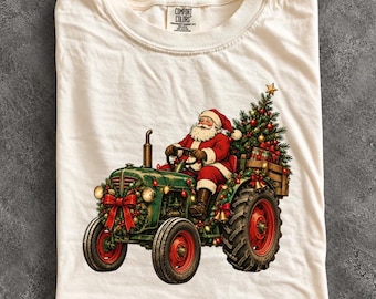 May include: An ivory t-shirt featuring a graphic of Santa Claus driving a green tractor decorated with Christmas lights and a red bow. The back of the tractor carries a Christmas tree with ornaments. The shirt has a Comfort Colors brand label.