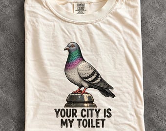 May include: An ivory t-shirt featuring a graphic of a pigeon perched on a post. The bird has grey, black, and iridescent purple-green neck feathers with red feet. Below the pigeon, the text reads: YOUR CITY IS MY TOILET. The shirt has a Comfort Colors label.