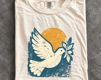 May include: An ivory t-shirt featuring a graphic print of a white dove carrying an olive branch. The design includes a large yellow sun set against a distressed blue rectangular background. The shirt has a Comfort Colors brand label at the neck.