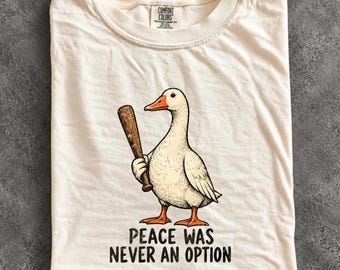 May include: An ivory t-shirt featuring a graphic of a white goose holding a brown baseball bat. Below the goose, the text reads, "PEACE WAS NEVER AN OPTION." The shirt has a Comfort Colors brand label at the inner neckline, indicating it is made of 100% cotton.