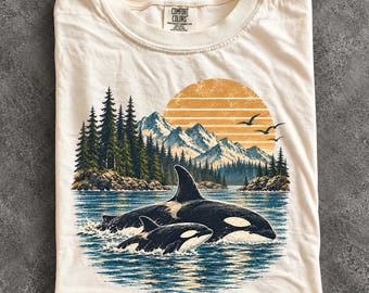 May include: An ivory t-shirt featuring a graphic print of two orcas swimming in blue water. The background shows a forest of green pine trees, snow-capped mountains, and a large orange sun with horizontal stripes. Three birds fly in the sky.