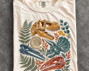 May include: An ivory t-shirt featuring a graphic print of four dinosaur skulls in yellow, blue, teal, and red, surrounded by green fern leaves and botanical elements. The inner neck label displays the text Comfort Colors.