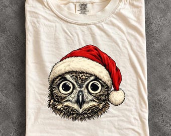 May include: An ivory t-shirt featuring a graphic of an owl wearing a red and white Santa hat. The owl has large black eyes and detailed brown and black feathers. The shirt has a Comfort Colors brand label printed on the inside neck area.