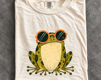 May include: An ivory t-shirt featuring a graphic of a green frog wearing orange-rimmed sunglasses. The frog has a light yellow belly and dark green spots. The shirt has a Comfort Colors brand label at the neck and the word IVORY at the bottom.