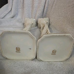 May include: A pair of off-white Lenox candle holders. Each holder features a rectangular base with gold trim and a detailed, twisted design. The Lenox logo is visible on the base. Made in the USA.