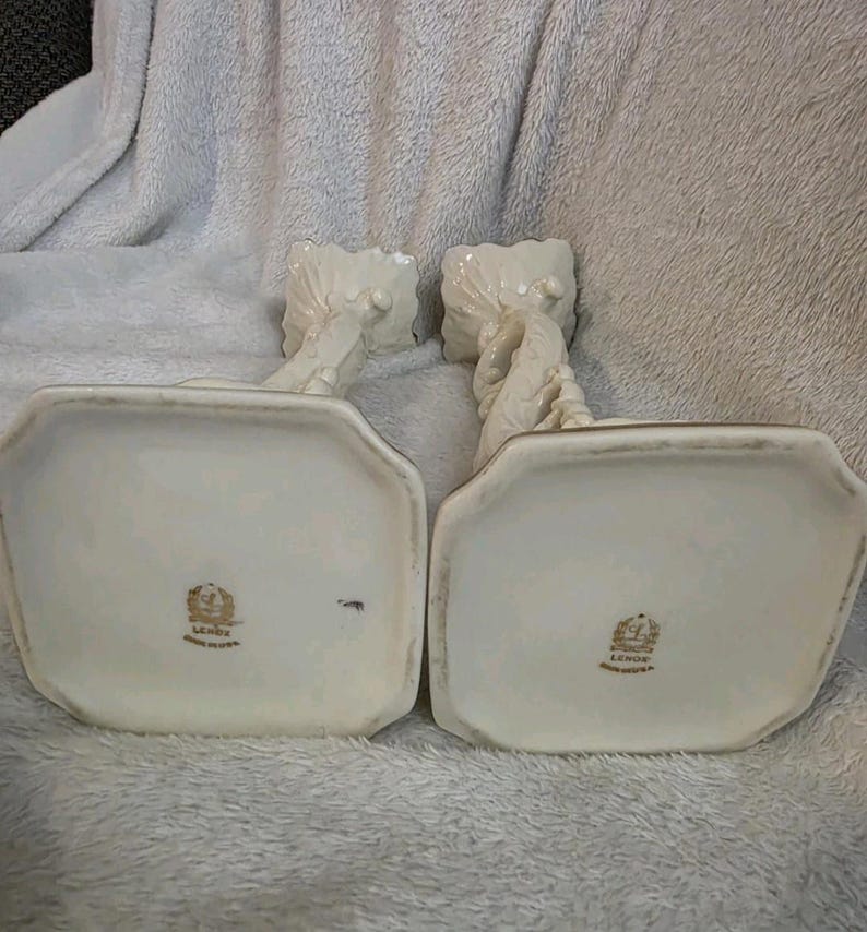 May include: A pair of off-white Lenox candle holders. Each holder features a rectangular base with gold trim and a detailed, twisted design. The Lenox logo is visible on the base. Made in the USA.