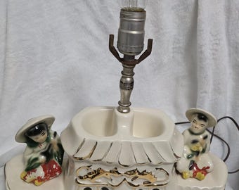 May include: A vintage ceramic lamp with a cream base, featuring a pagoda design with gold detailing. Two figurines in traditional dress are positioned on either side. The lamp has a silver metal stem and a clear lightbulb.
