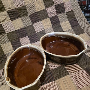 Puede incluir: Dos fuentes de horno ovaladas de cerámica marrón con acabado brillante. Las fuentes tienen un borde blanquecino y están sobre un edredón de retazos con cuadrados marrones, beige y crema.