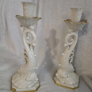 May include: A pair of white ceramic candle holders with a dolphin-inspired design. Each candle holder has a scalloped top and base, both trimmed with gold. The stems feature a curved, ornate design. The candle holders are displayed on a white surface.