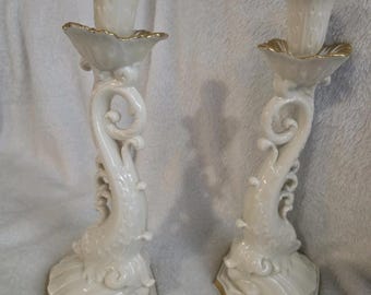 May include: Two white ceramic candle holders with a dolphin-like design. The candle holders have a scalloped top and base with gold trim. The stems feature a curved, ornate design. The candle holders are displayed on a white surface.