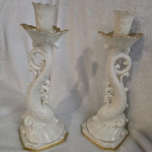 May include: A pair of white ceramic candlesticks with gold detailing. The candlesticks feature a dolphin-inspired design with a flared base and a decorative, scalloped top. Gold trim highlights the edges of the base and the top.