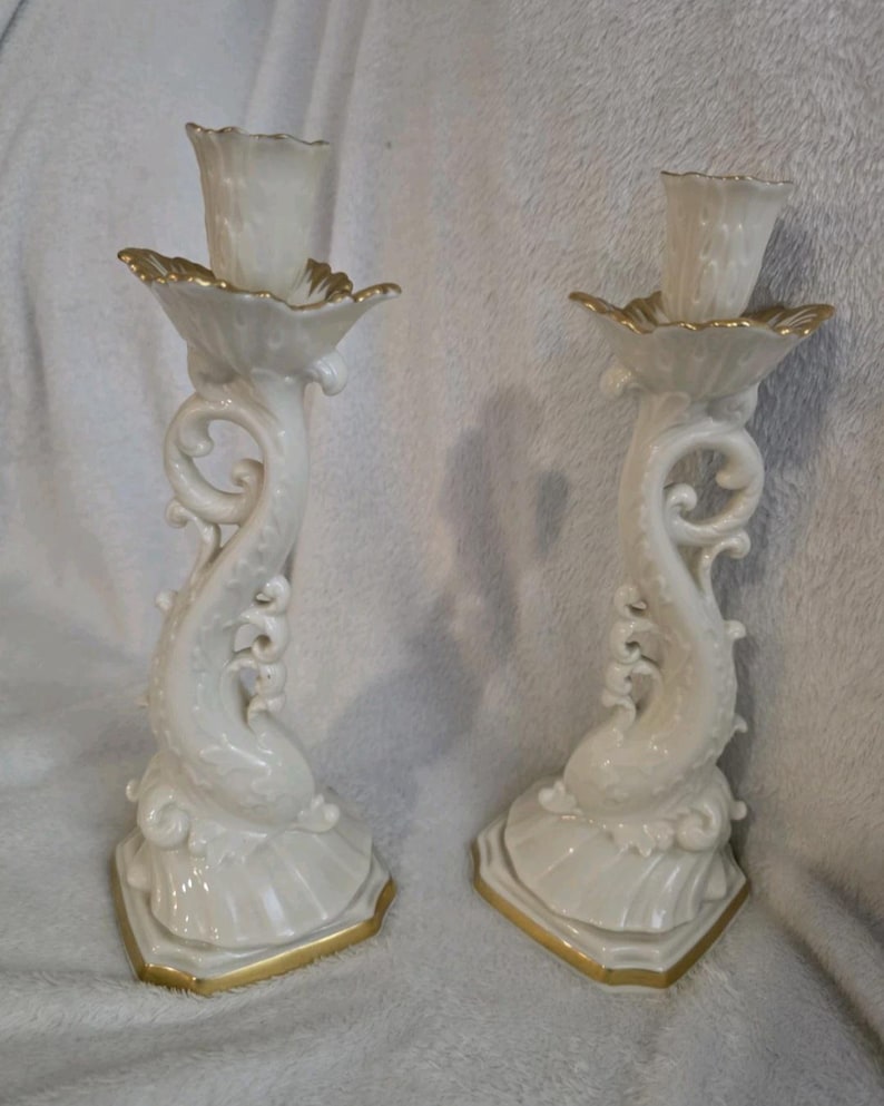 May include: A pair of white ceramic candlesticks with gold detailing. The candlesticks feature a dolphin-inspired design with a flared base and a decorative, scalloped top. Gold trim highlights the edges of the base and the top.