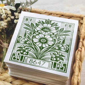 May include: A stack of square white paper napkins featuring a dark green printed design of stylized flowers and leaves framed by geometric borders. The number 8647 is printed in a rectangular box at the bottom of the green floral illustration.