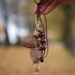 May include: A handmade keychain featuring a cluster of natural, textured brown seed pods and wooden beads. The keychain includes a leather cord, a metal clasp, and the text "Sylvia & Sage" in the upper right corner.