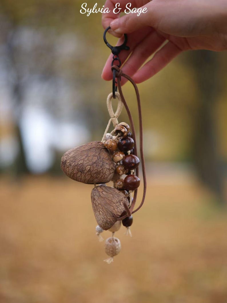 May include: A handmade keychain featuring a cluster of natural, textured brown seed pods and wooden beads. The keychain includes a leather cord, a metal clasp, and the text "Sylvia & Sage" in the upper right corner.
