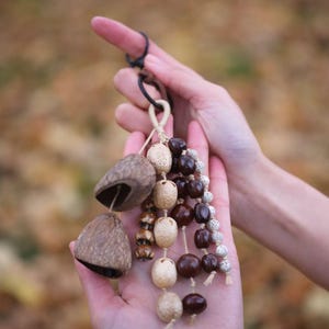 May include: A handcrafted, natural-toned ornament featuring two bell-shaped seed pods and strands of various beads in shades of brown, beige, and white. The ornament is held in open hands against a blurred, autumnal background. The text "Sylvia & Sage" is in the upper left.