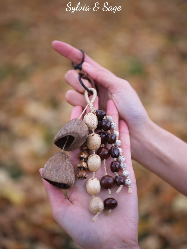 May include: A handcrafted, natural-toned ornament featuring two bell-shaped seed pods and strands of various beads in shades of brown, beige, and white. The ornament is held in open hands against a blurred, autumnal background. The text "Sylvia & Sage" is in the upper left.