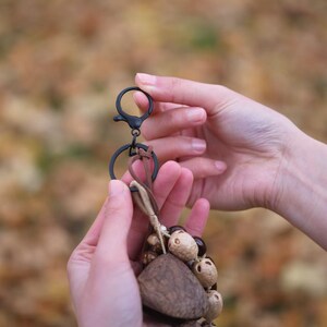 May include: A keychain with a black clasp and ring, featuring a cluster of natural-toned wooden and seed-like beads. The keychain is held in two hands against a blurred, autumnal background. The text "Sylvia & Sage" is at the top.