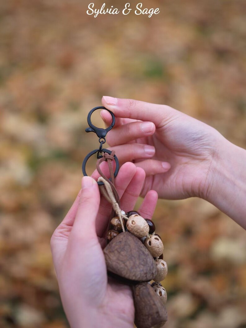 May include: A keychain with a black clasp and ring, featuring a cluster of natural-toned wooden and seed-like beads. The keychain is held in two hands against a blurred, autumnal background. The text "Sylvia & Sage" is at the top.