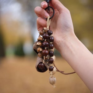 May include: A handmade keychain featuring natural elements. The keychain includes a black metal clasp, a tan cord, and various brown and tan beads and pods. The text "Sylvia & Sage" is in the upper left corner.