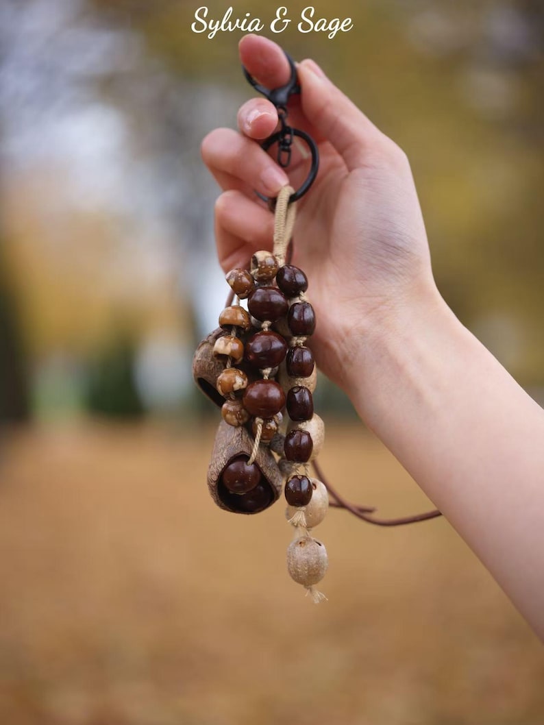 May include: A handmade keychain featuring natural elements. The keychain includes a black metal clasp, a tan cord, and various brown and tan beads and pods. The text "Sylvia & Sage" is in the upper left corner.