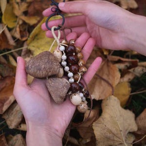 May include: A handmade keychain featuring natural elements. The keychain includes a black metal clasp, wooden beads, and three large, textured, mushroom-shaped charms. The background is a bed of autumn leaves. The text "Sylvia & Sage" is in the upper left corner.