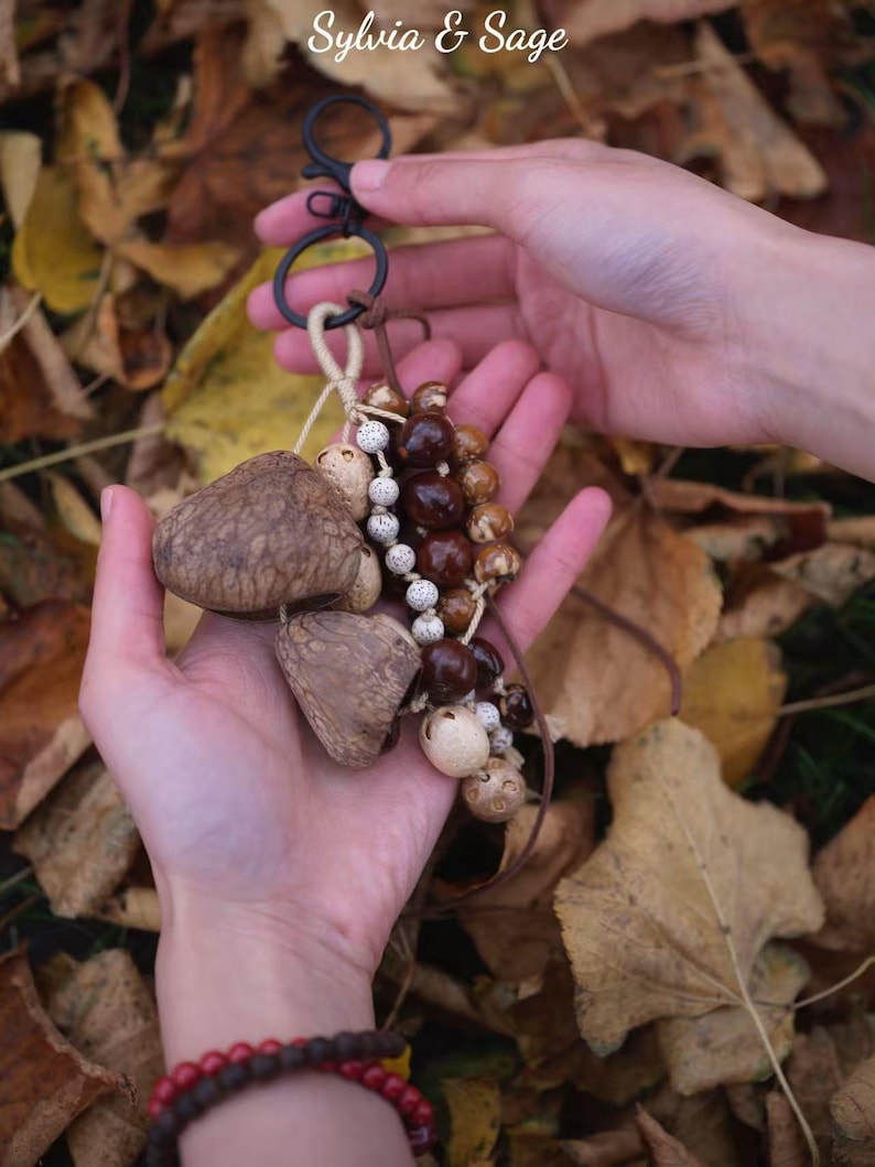 May include: A handmade keychain featuring natural elements. The keychain includes a black metal clasp, wooden beads, and three large, textured, mushroom-shaped charms. The background is a bed of autumn leaves. The text "Sylvia & Sage" is in the upper left corner.