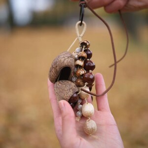 May include: A handmade pendant featuring natural elements. The pendant includes a cluster of brown and tan nuts and seeds, suspended from a leather cord. The text "Sylvia & Sage" is visible in the upper left corner.