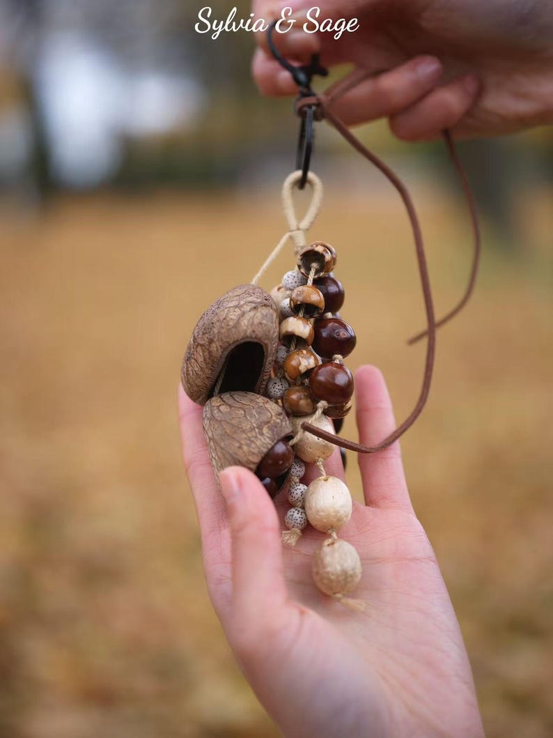 May include: A handmade pendant featuring natural elements. The pendant includes a cluster of brown and tan nuts and seeds, suspended from a leather cord. The text "Sylvia & Sage" is visible in the upper left corner.