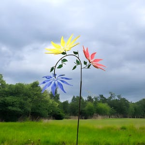 Puede incluir: Una estaca de jardín colorida con tres giradores en forma de flor en amarillo, rosa y azul. Las flores están unidas a un tallo de metal negro curvado con detalles de hojas verdes. El arte del jardín se encuentra sobre un fondo de hierba verde y árboles.