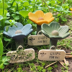 Puede incluir: Tres bebederos para abejas con forma de flor de colores en un jardín. Las flores son amarillas, azules y verdes, cada una con un centro texturizado. Pequeños letreros de madera dicen "CUPS FOR BEE" y presentan una ilustración de abeja.