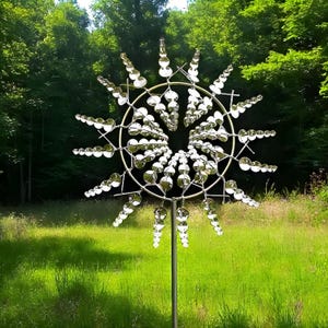 May include: A silver metal kinetic wind spinner with a circular design and multiple reflective elements. The outdoor garden art is set against a backdrop of green grass and trees, creating a dynamic visual effect.