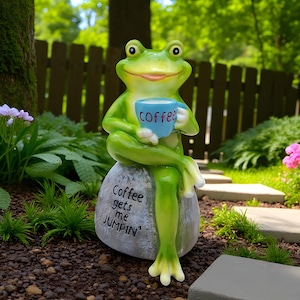 Puede incluir: Una estatua de rana verde caprichosa sosteniendo una taza de café azul con la palabra "coffee". La rana está sentada en una piedra gris con las palabras "Coffee gets me JUMPIN'". La estatua está en un jardín.