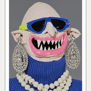 Könnte beinhalten: Eine skurrile Skulptur eines Gesichts mit blauer Sonnenbrille mit gelbem Akzent, einem rosa Mund mit weißen Zähnen und großen diamantartigen Ohrringen. Das Gesicht trägt einen blauen Rollkragenpullover und eine weiße Perlenkette.