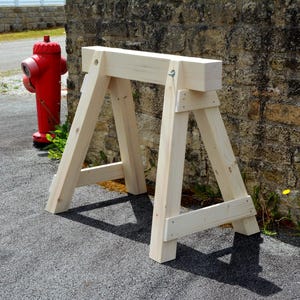 Könnte beinhalten: Eine hellfarbene Holz-Sägebock steht auf einer grauen Asphaltfläche. Der Sägebock hat abgewinkelte Beine und einen horizontalen oberen Balken. Ein roter Hydrant ist im Hintergrund zu sehen, zusammen mit einer Steinmauer.