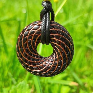 May include: A black and copper-colored pendant necklace. The pendant is a circular shape with a woven design. The necklace cord is black and knotted, with a gold-colored bead. The background is blurred green foliage and yellow flowers.
