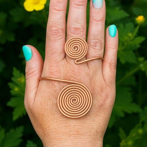 Könnte beinhalten: Eine Hand zeigt Kupferschmuck: einen Spiralring, einen Spiral-Fingerring und ein geflochtenes Armband. Die Hand hat türkisfarbenen Nagellack. Der Hintergrund zeigt grünes Laub und gelbe Blumen.