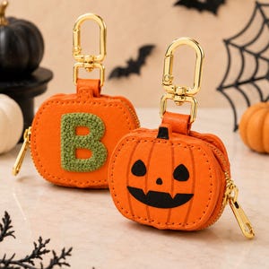 May include: Two orange pumpkin-shaped zipper pouches with gold-tone metal lobster clasps. One pouch features a green textured letter B, while the other displays a black jack-o'-lantern face with triangular eyes and a wide, jagged grin.