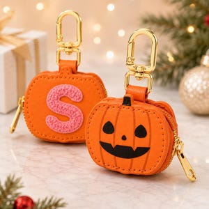 May include: Two orange pumpkin-shaped zip pouches with gold-tone metal lobster claw clasps. One pouch features a textured pink letter S patch, while the other displays a black jack-o'-lantern face with triangular eyes and a jagged mouth.