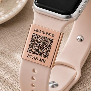 Puede incluir: Una correa de reloj inteligente de silicona rosa claro con un dije de metal en oro rosa grabado con un código QR, el texto "HEALTH INFOR" arriba del código y "SCAN ME" debajo. El reloj tiene una caja plateada, pantalla negra y corona.