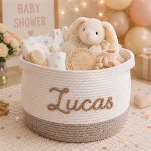 Op de afbeelding: Een witte touwmand met een bruine basis en de naam "Lucas" in bruin schrift. De mand is gevuld met babyartikelen, waaronder een knuffelkonijn, kleding, een houten bord met de tekst "hello LITTLE ONE" en een borstel. Een "BABY SHOWER" bord staat op de achtergrond.