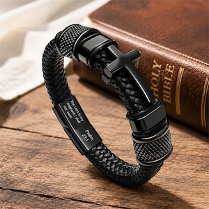 Pode incluir: Uma pulseira trançada preta com um pingente de cruz preta central e um fecho metálico retangular gravado com o texto The Lord is my shepherd, I shall not want e Psalm 23:1. A pulseira inclui detalhes em metal preto liso.