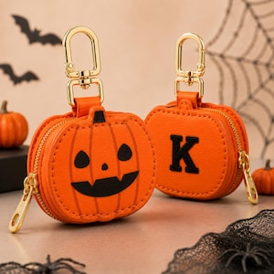 May include: Two orange pumpkin-shaped pouches with gold-tone metal lobster clasps and zippers. One pouch features a black jack-o'-lantern face, while the other displays a black embroidered letter K. Both items are round with stitched vertical lines.