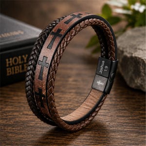May include: A multi-strand bracelet featuring a central brown leather band with black cross symbols and two flanking braided dark brown leather cords. The bracelet includes a black metal clasp engraved with the text Joshua 1:9 and a small cross.