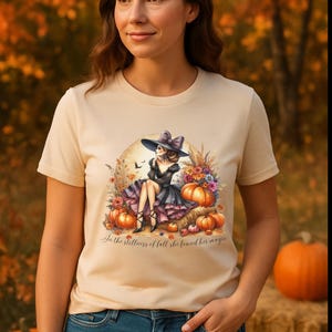 May include: Beige t-shirt featuring a whimsical illustration of a witch in a black dress and hat, surrounded by pumpkins and flowers. The text "In the stillness of fall she found her magic" is below the illustration.