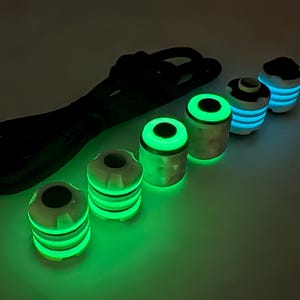 Könnte beinhalten: Eine Sammlung leuchtender, zylindrischer Objekte und ein schwarzes Kabel. Die Objekte haben eine weiße Oberseite und einen silbernen Körper mit hellgrünen und blauen Leuchtringen. Das Kabel ist schwarz und scheint leicht zu leuchten.