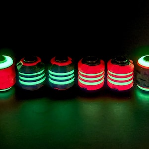 Peut inclure: Six perles cylindriques disposées en rangée sur un fond sombre. Chaque perle présente un motif distinct de bandes lumineuses vert fluo ou une illumination rouge solide, certaines affichant une combinaison des deux couleurs vives en surface.